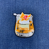 An embroidered patch of a whiskey old fashioned cocktail, placed on a blue denim fabric background.