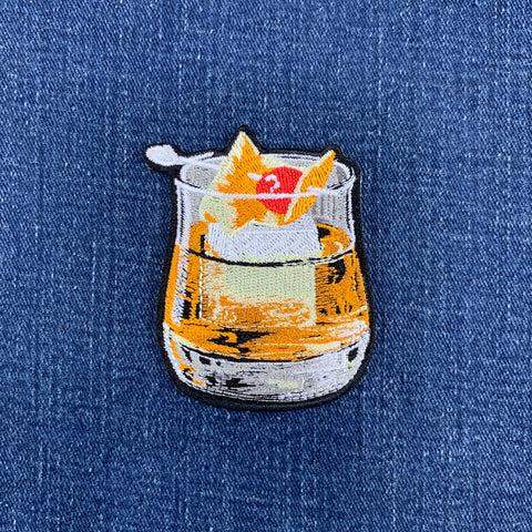 An embroidered patch of a whiskey old fashioned cocktail, placed on a blue denim fabric background.