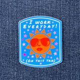 A sticker with a tan background and a cartoon sun character wearing sunglasses. The text on the sticker reads 'I WORK EVERYDAY (ON THIS TAN)' surrounded by pink stars against a blue background.