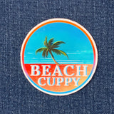 A round multicolored sticker with a palm tree and the text 'BEACH CUPPY' on a denim fabric background.
