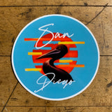 A circular sticker of a pelican is set against a striped orange and yellow sunset on a bright blue background. The words 'San Diego' are written above and below the bird.