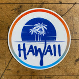 A circular sticker with a white background and a gradient border of orange, blue, and white. The center design features a dark blue sunset/ocean scene with the white silhouette of three palm trees, and the word 'HAWAII' written in large, brush-style dark blue letters across the bottom.