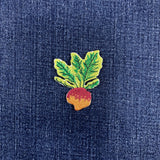 A small embroidered Rutabaga patch with green leaves and a red-orange root, placed on a textured dark blue fabric background.