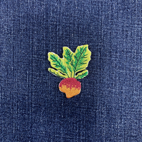 A small embroidered Rutabaga patch with green leaves and a red-orange root, placed on a textured dark blue fabric background.