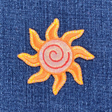 A sparkly embroidered sun applique patch with yellow and orange rays and a golden spiral center on a denim background