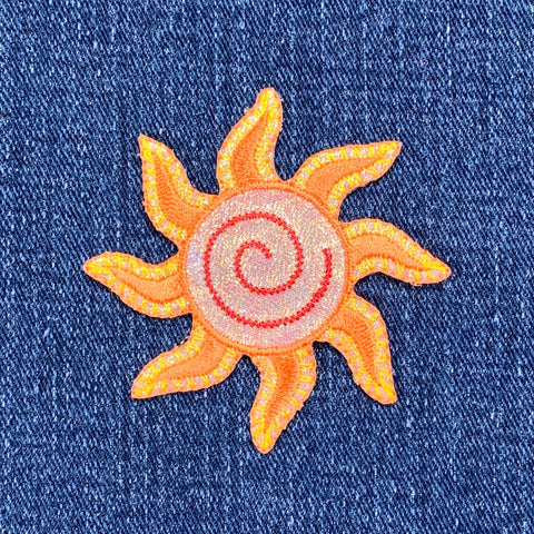 A sparkly embroidered sun applique patch with yellow and orange rays and a golden spiral center on a denim background