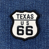 Embroidered patch in the shape of a shield with 'TEXAS US 66' text, primarily black and white, and displayed on a denim background