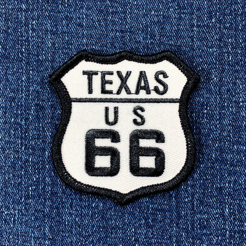 Embroidered patch in the shape of a shield with 'TEXAS US 66' text, primarily black and white, and displayed on a denim background