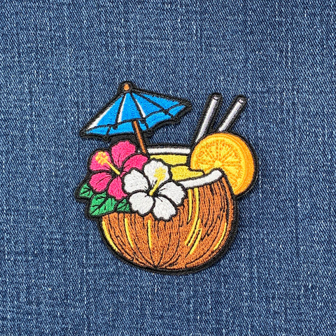 An embroidered patch of a tropical coconut cocktail with a blue umbrella, pink and white flowers, and an orange slice, shown on a blue denim fabric background.