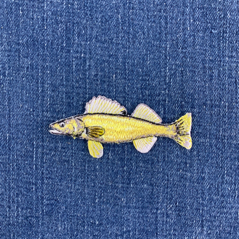 A small embroidered walleye fish patch in yellow and white, placed on a blue denim fabric background.