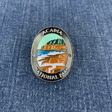 An oval enamel Walking Stick Medallion featuring a landscape of Acadia National Park with a castle, mountains, and blue water, bordered by a silver metal frame with the text 'ACADIA NATIONAL PARK' on a blue denim background.
