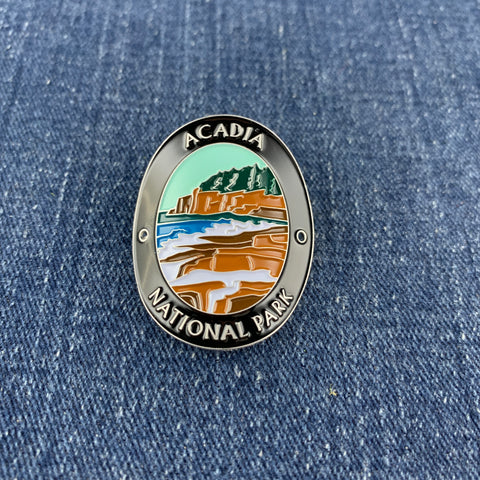 An oval enamel Walking Stick Medallion featuring a landscape of Acadia National Park with a castle, mountains, and blue water, bordered by a silver metal frame with the text 'ACADIA NATIONAL PARK' on a blue denim background.
