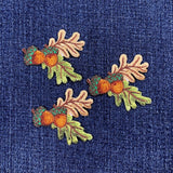 Three embroidered patches featuring acorns and oak leaves in green and brown colors, designed to be ironed onto fabric, and displayed on a denim background