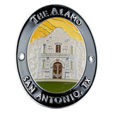 An oval medallion featuring an illustrated view of The Alamo in San Antonio, TX. The design has a black outer border with raised silver text reading 'THE ALAMO' at the top and 'SAN ANTONIO, TX' at the bottom, a silver inner ring, and a central multicolor illustration of the Alamo building with a yellow sky and green ground.