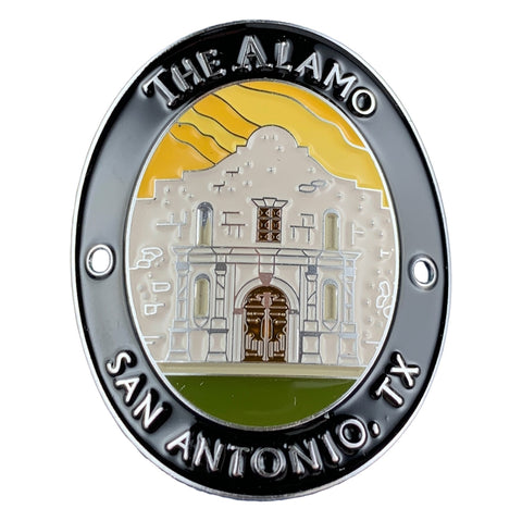 An oval medallion featuring an illustrated view of The Alamo in San Antonio, TX. The design has a black outer border with raised silver text reading 'THE ALAMO' at the top and 'SAN ANTONIO, TX' at the bottom, a silver inner ring, and a central multicolor illustration of the Alamo building with a yellow sky and green ground.