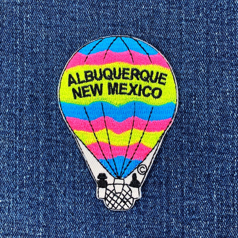 Embroidered patch of a hot air balloon with bright neon stripes (blue, pink, yellow) reading "ALBUQUERQUE NEW MEXICO" against a denim background.