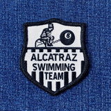 A shield-shaped patch with the text 'Alcatraz Swimming Team' and an illustration of a figure in a straight jacket and an 8-ball.