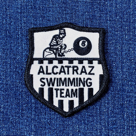 A shield-shaped patch with the text 'Alcatraz Swimming Team' and an illustration of a figure in a straight jacket and an 8-ball.