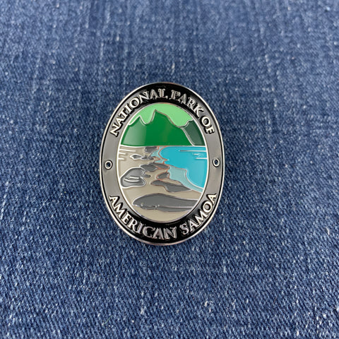 An oval metal enamel Walking Stick Medallion featuring the National Park of American Samoa emblem, resting on a blue denim fabric background.