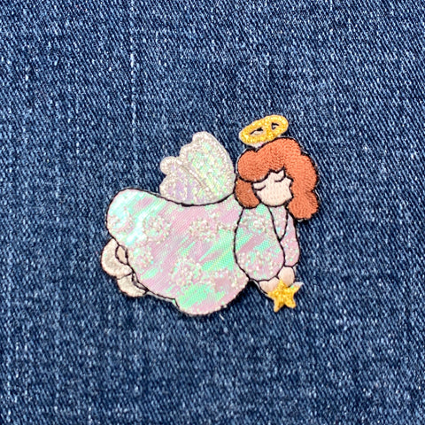 An embroidered patch depicting an angel with a halo, praying with a star in hand, finished with a shimmery finish, displayed on a denim background