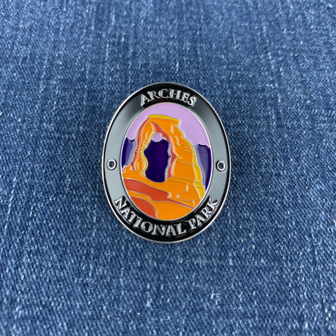 An oval enamel Walking Stick Medallion featuring a desert arch landscape with the text 'ARCHES NATIONAL PARK' on a blue denim background.