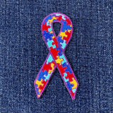 A ribbon-shaped patch with a puzzle piece design, featuring colors red, blue,  and yellow, with a purple outline, representing autism awareness.