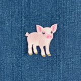 A small embroidered pig patch in light pink with darker pink inner ears and snout, black eyes, and tiny gold-toned hooves, placed on a textured blue fabric background.