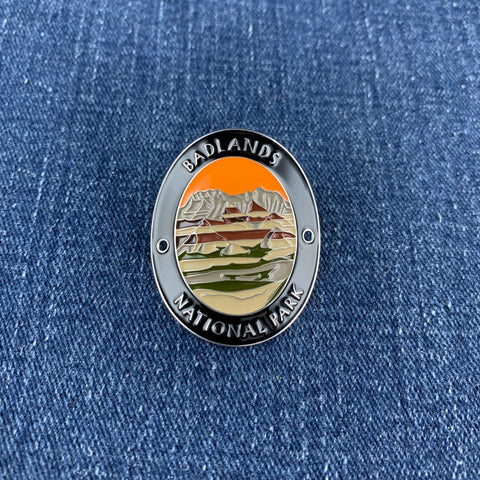 An oval enamel medallion featuring the Badlands National Park landscape, displayed on a blue denim fabric background.