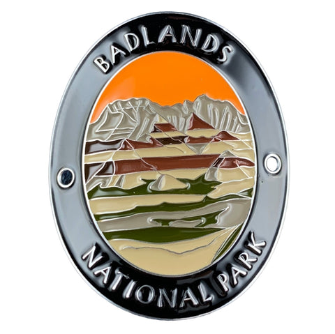 An oval enamel medallion featuring a landscape design of Badlands National Park. The pin has a polished silver-tone border with raised text reading 'BADLANDS NATIONAL PARK' at the top and bottom.