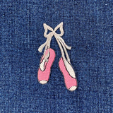 A pair of pink ballet slippers with silver accents, tied with a ribbon, designed as an iron-on applique patch, displayed on a denim background