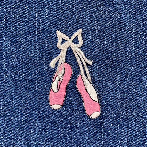 A pair of pink ballet slippers with silver accents, tied with a ribbon, designed as an iron-on applique patch, displayed on a denim background