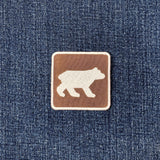 A square brown embroidered patch with a white bear silhouette, shown on a blue denim fabric background.