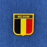 A shield-shaped Belgium patch with black, yellow, and red vertical stripes and the word BELGIUM in yellow, shown on a blue textured fabric background.