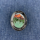 An oval enamel Walking Stick Medallion featuring a landscape of Big Bend National Park with orange mountains, green hills, and white grass, bordered by a black ring with white lettering, shown on a blue denim background.