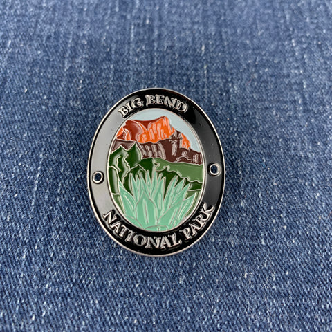 An oval enamel Walking Stick Medallion featuring a landscape of Big Bend National Park with orange mountains, green hills, and white grass, bordered by a black ring with white lettering, shown on a blue denim background.