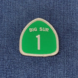 A green shield-shaped embroidered patch with a white border, featuring the text 'BIG SUR' and the number '1' in cream, placed on a blue denim background.