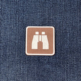 A square brown patch with a white binocular icon, placed on a blue denim fabric background.