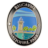 An oval Biscayne National Park enamel medallion featuring a silver-toned border with raised lettering and a colorful central illustration of a lighthouse, palm trees, water, and sky.