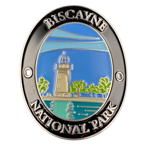An oval Biscayne National Park enamel medallion featuring a silver-toned border with raised lettering and a colorful central illustration of a lighthouse, palm trees, water, and sky.