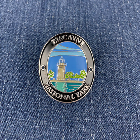 An oval metal enamel Walking Stick Medallion featuring the Biscayne National Park lighthouse design, placed on a blue denim background.