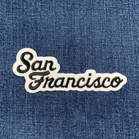 A black and white embroidered patch with "San Francisco" in a cursive script, set against a background of blue denim fabric.