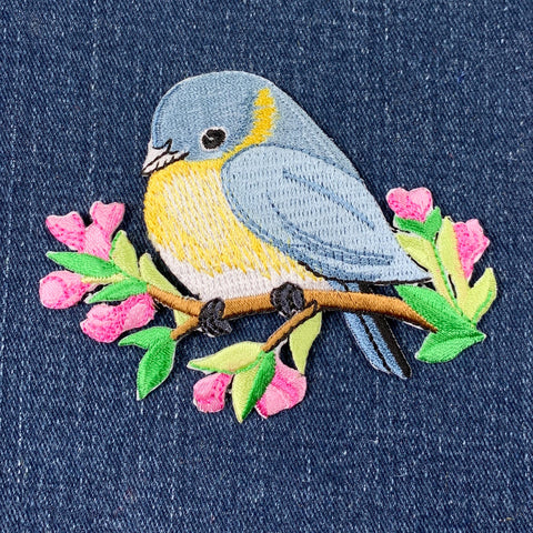 Embroidered patch of a blue and yellow bird on a branch with pink flowers on denim fabric.