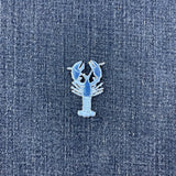 A small lobster-shaped enamel patch in light blue and white, placed on a textured blue denim fabric background.