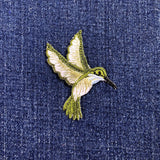 An embroidered hummingbird patch in olive green and cream, shown on a textured dark blue denim fabric background.