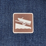 A square embroidered patch featuring a white boat and trailer graphic on a brown background, placed on a blue denim fabric surface.