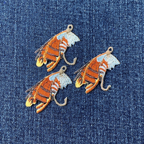 Three embroidered patches depicting fly fishing lures with hooks, wings, and detailed patterns on a denim background