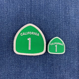 Two green California Route 1 road sign patches with white borders and white lettering, displayed on a blue denim fabric background.