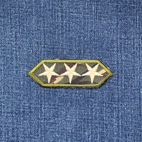 A patch featuring three white stars on a camouflage fabric, displayed on a denim background.