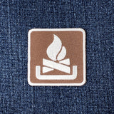 A square embroidered patch featuring a white campfire logo on a brown background, placed on a blue denim fabric surface.