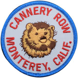 A circular embroidered patch featuring a brown and tan sea otter in the center, surrounded by red text reading 'CANNERY ROW MONTEREY, CALIF.' on a white background with a blue stitched border.